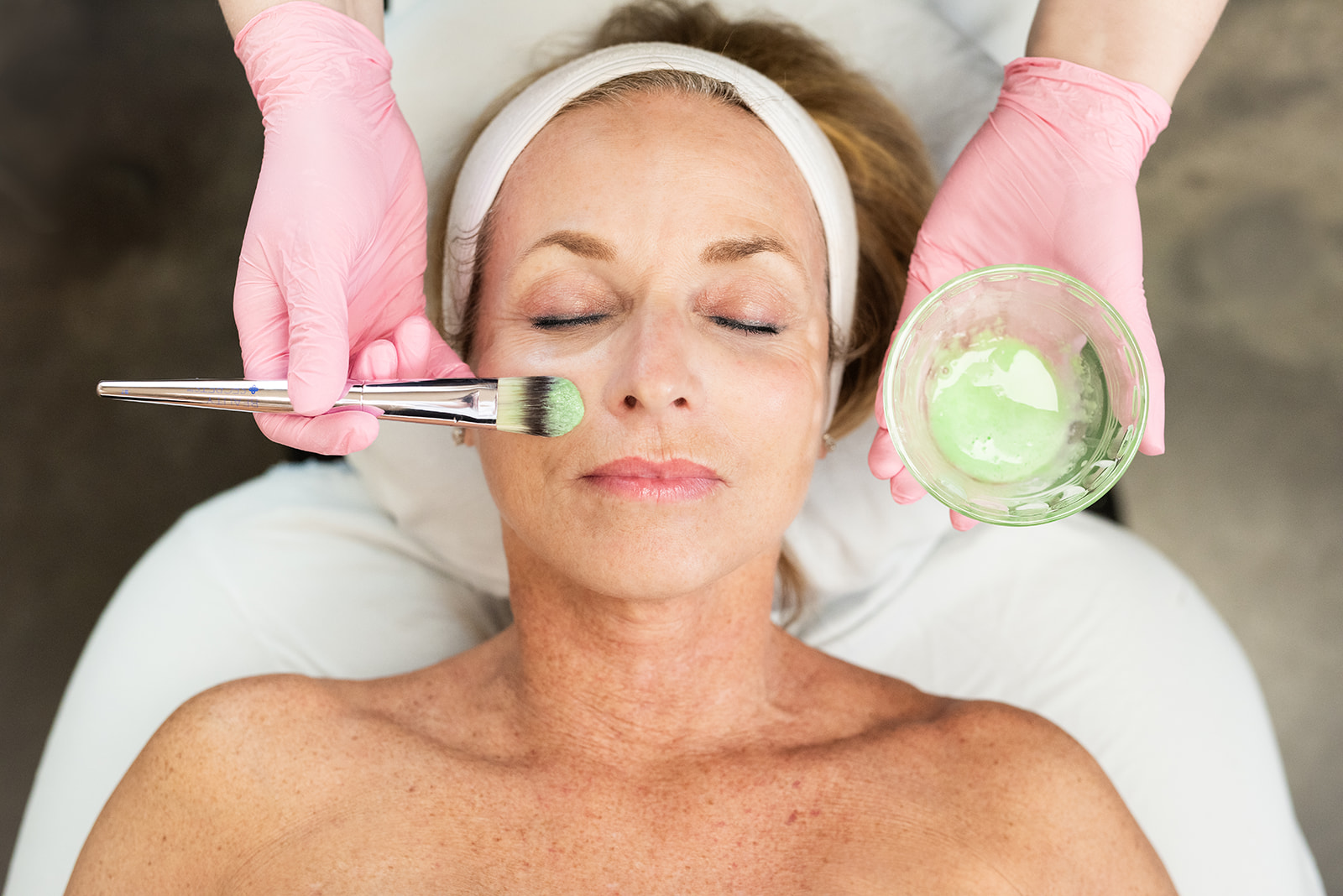 Woman receiving a facial treatment at Kamloops Skin Solutions while an aesthetician applies a green mask with a brush.